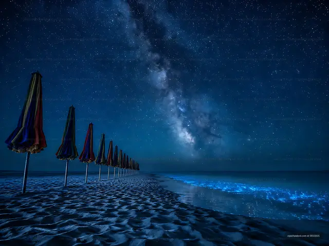 Night sky over a beach with umbrellas and glowing ocean waves